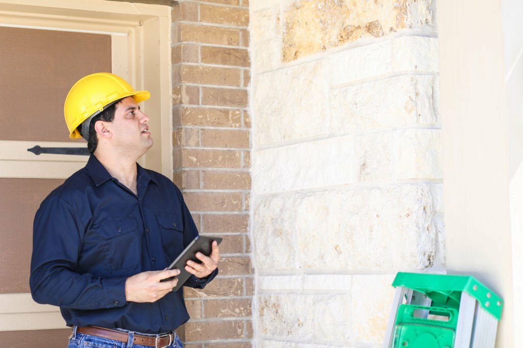 Latin descent man inspects home exterior. He could be a home inspector, insurance adjuster, exterminator or a variety of blue collar occupations. He holds a digital tablet, wears hard hat and blue work shirt.